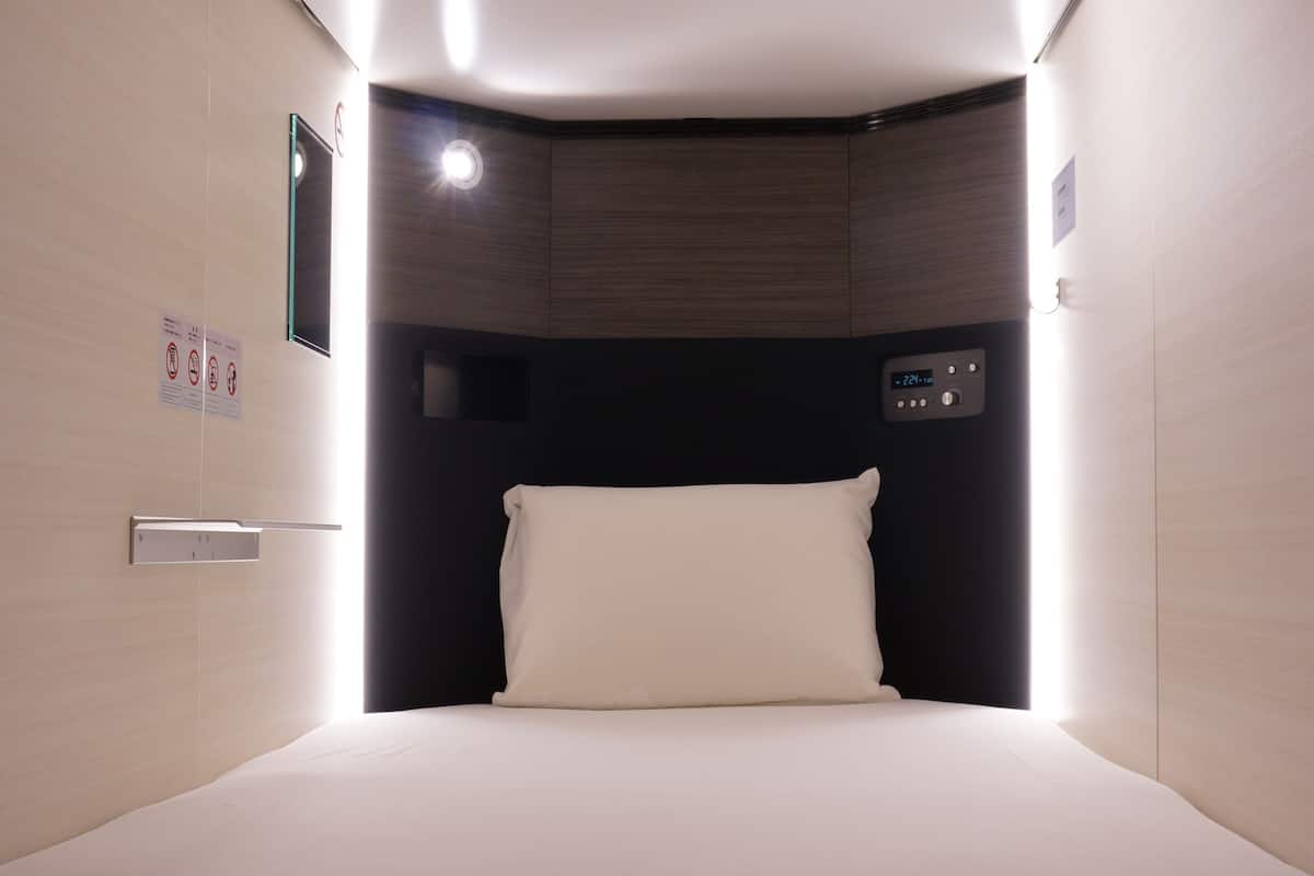 Una habitación de hotel con cama, panel de control y una lámpara fijada a la pared.