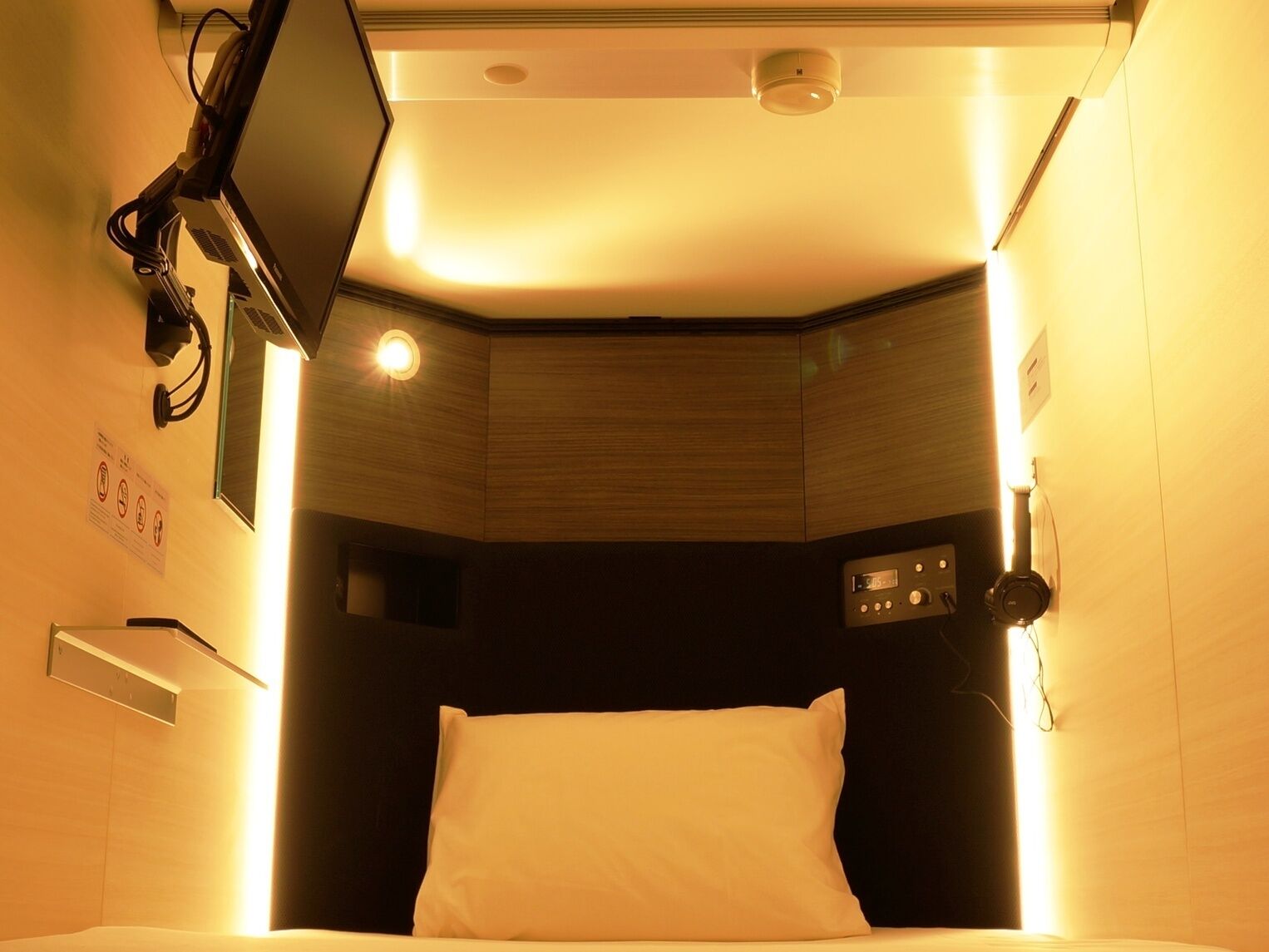 A compact hotel room with a bed, a mounted television, a control panel, and a ceiling-mounted air conditioning unit.
