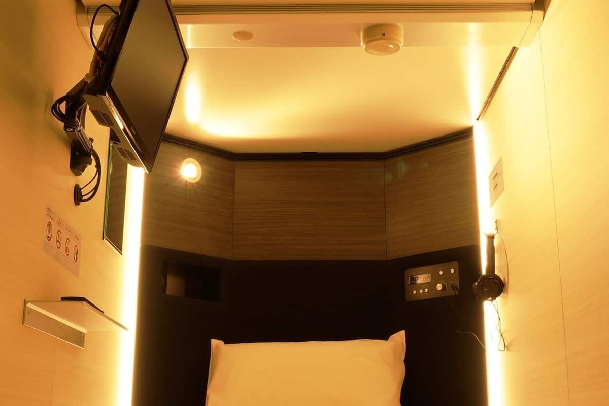 Habitación de hotel compacta con cama, televisor montado, panel de control y aire acondicionado instalado en el techo.