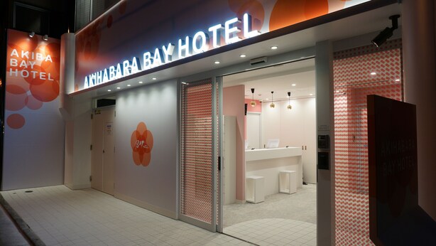 Exterior del Hotel Akihabara Bay, con un diseño moderno, que presenta una zona de recepción con mostrador y asientos, y una entrada visible.