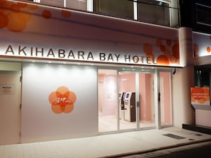 The exterior of Akihabara Bay Hotel with a prominent sign, a glass entrance, and a visible vending machine.