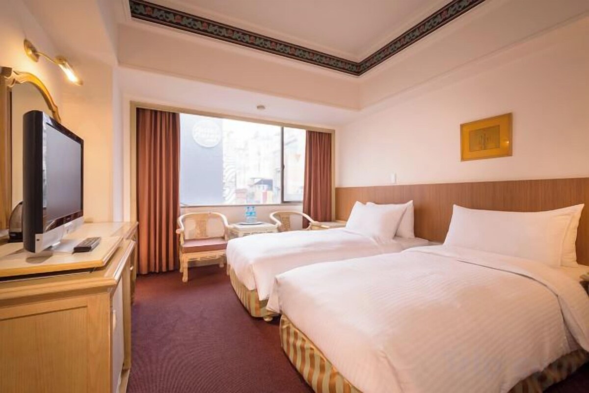 A hotel room with two beds, a desk with a television, a chair, and a window with curtains.