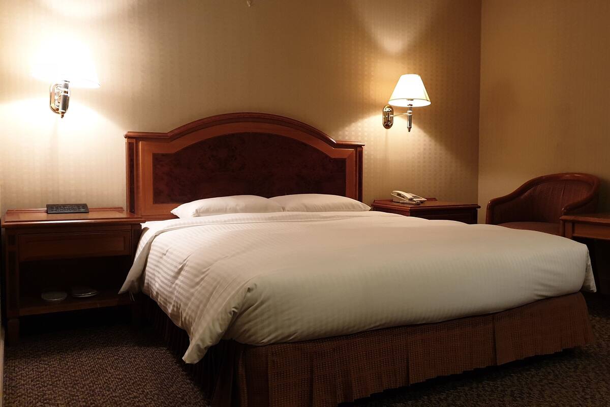 A hotel room with a large bed, wooden headboard, two bedside tables with lamps, a telephone, and a chair.