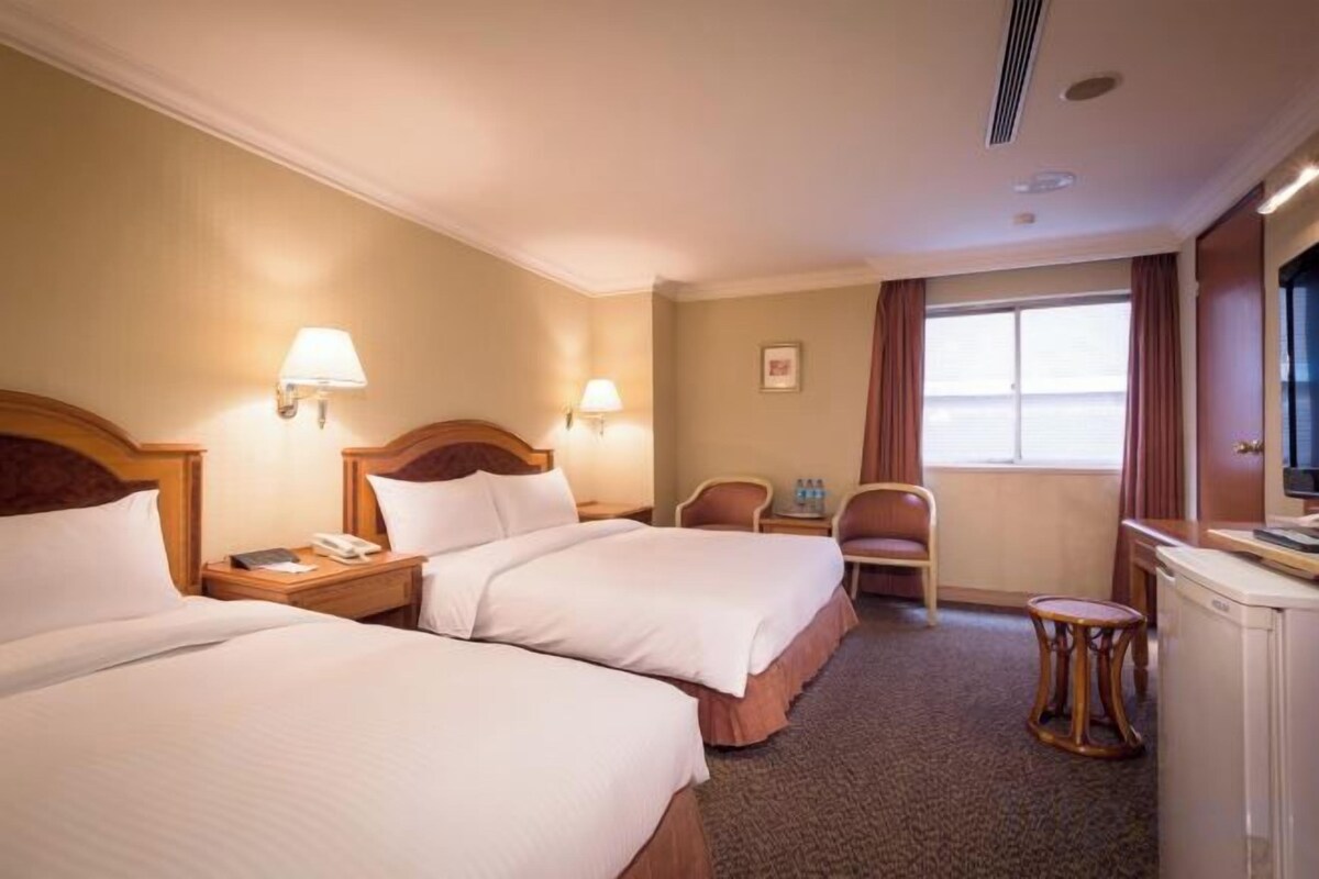 A hotel room with two beds, a desk, a chair, a television, and a window with curtains.