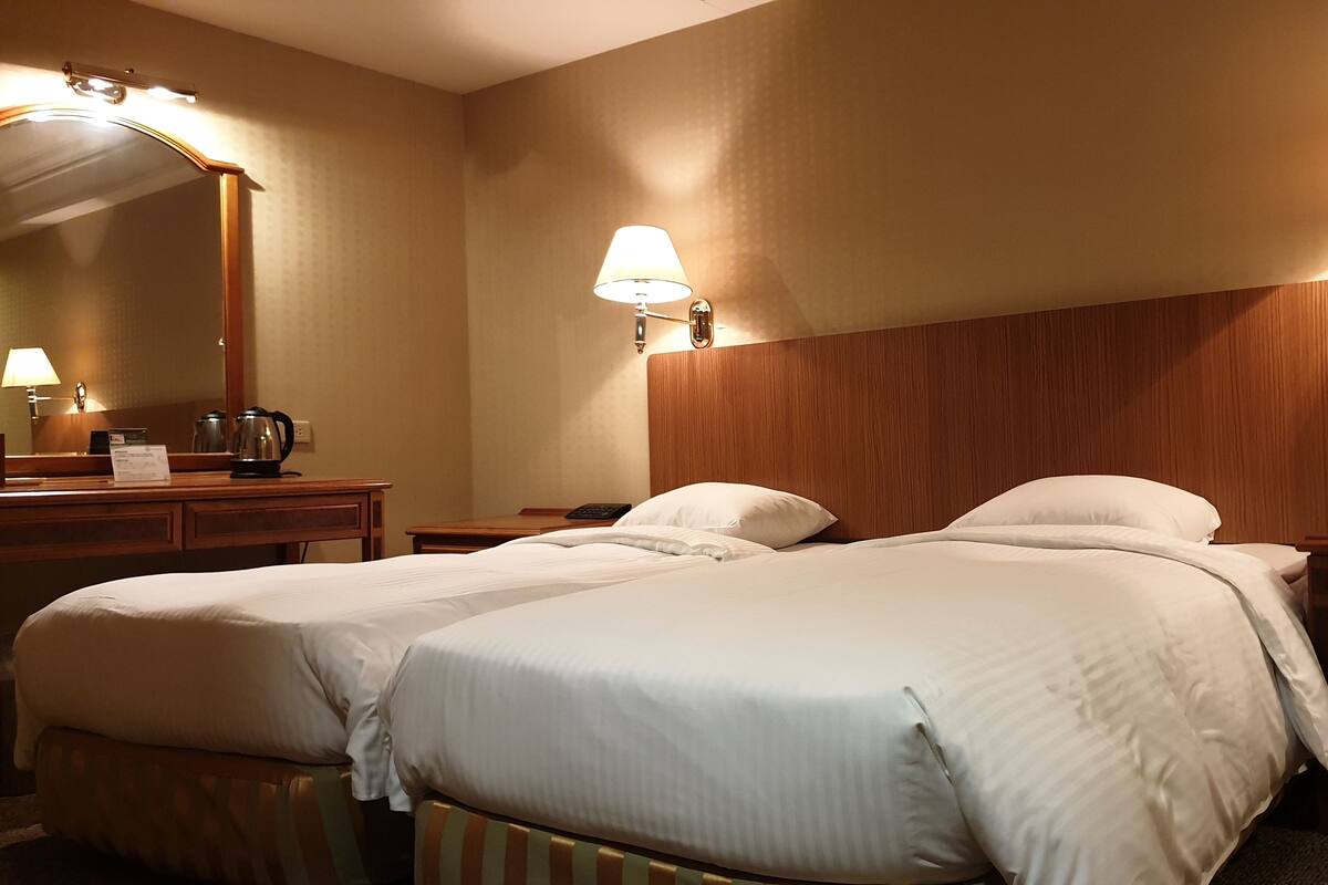 A hotel room with two beds, a wooden headboard, a desk with a lamp, a telephone, and a mirror.