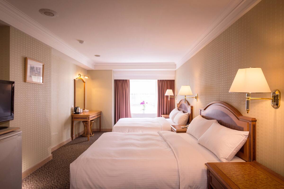 A hotel room with two beds, a television, a desk with a lamp, and a window with curtains.