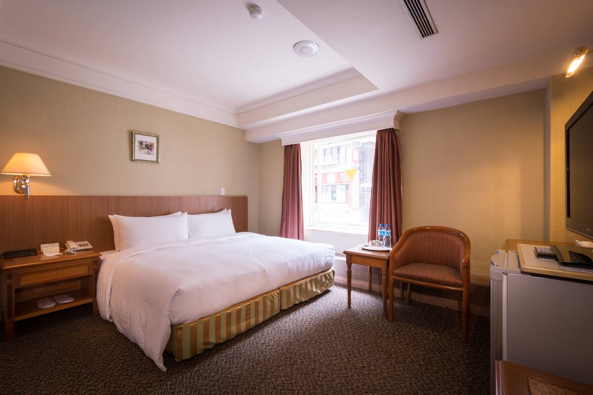 A hotel room with a large bed, a desk with a chair, a television, and a window with curtains.