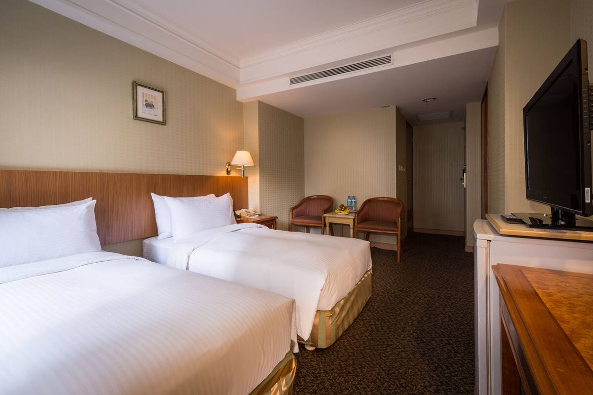 A hotel room with two beds, a desk, a chair, a television, and a lamp.