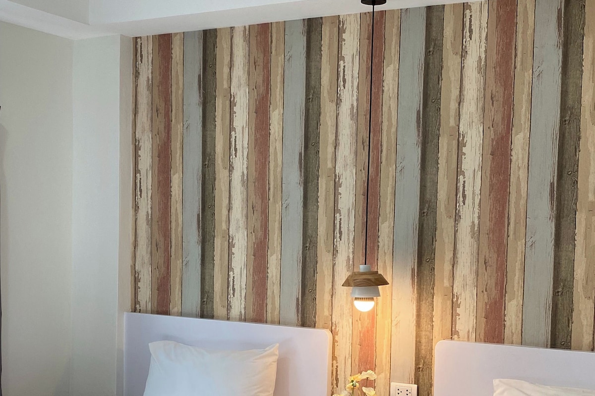 A hotel room with two beds, a wooden wall panel, a small table with a vase of flowers, and a hanging light fixture.
