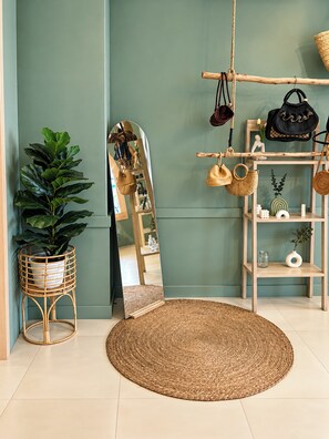 Une pièce avec un grand miroir, un tapis rond en fibres naturelles, une plante en pot et une étagère en bois où sont posés des sacs et des chapeaux.