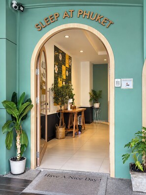 L’entrée d’un hôtel, avec une enseigne indiquant « DORMIR À PHUKET », un comptoir d’accueil, une plante en pot et un paillasson.