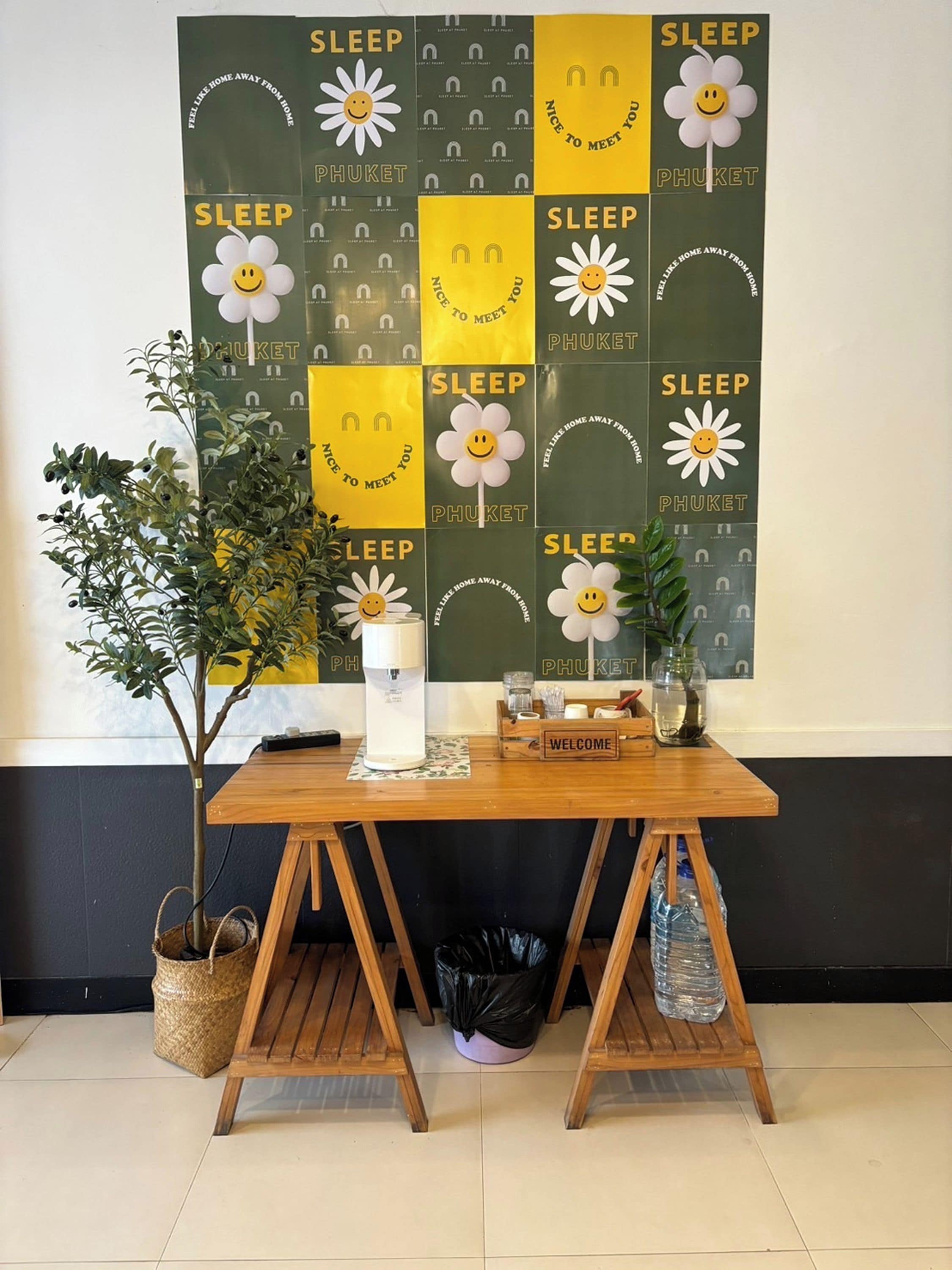 A room with a wooden table, a potted plant, a water bottle, a coffee machine, and a welcome sign.