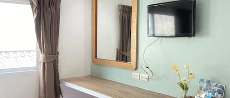 A hotel room with a bed, a desk with a flat-screen TV, a mirror, a window with curtains, and a wall-mounted basket with toiletries.