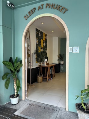 L'ingresso di un hotel chiamato 'Sleep at Phuket', con un cartello sopra la porta, una pianta in vaso a sinistra e un piccolo tavolo con una pianta all'interno.