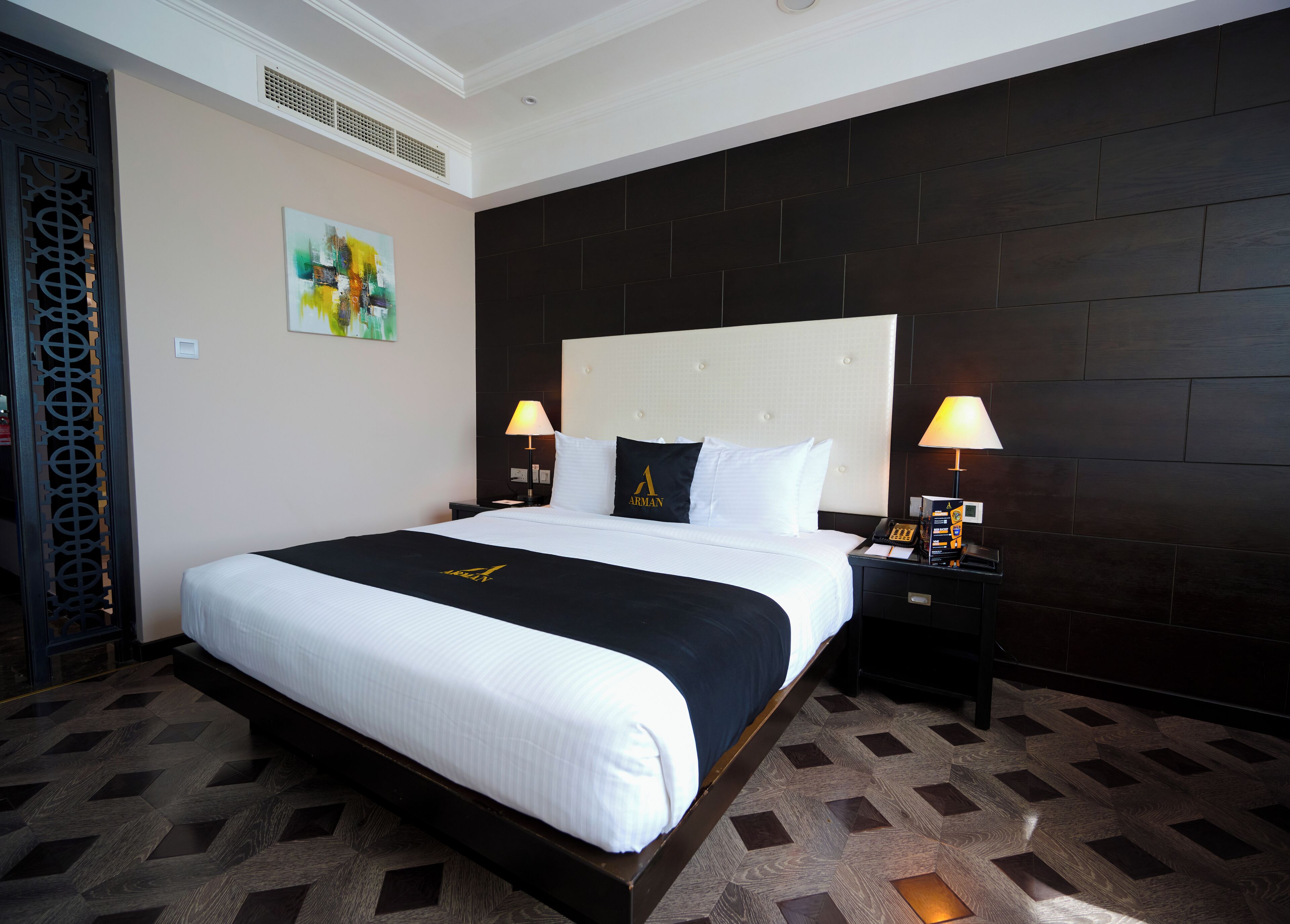 A hotel room with a large bed, two bedside tables with lamps, a decorative wall panel, and a painting.