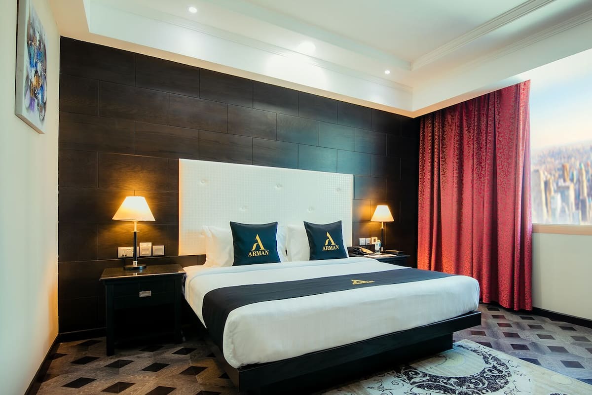 Camera d'albergo con un letto grande, due abat-jour, una testiera con un logo, un tappeto a motivi e vista sulla città attraverso una finestra con tende rosse.