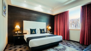 Habitación de hotel con una cama grande, dos lámparas de noche, un cabecero con un logotipo, una alfombra estampada y vistas de un paisaje urbano a través de una ventana con cortinas rojas.