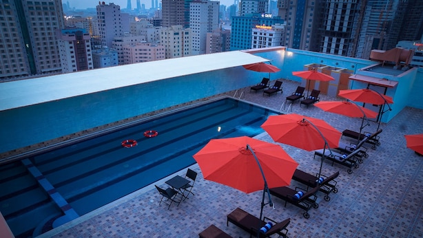 Un área de piscina en la azotea con sillones y sombrillas, rodeada de edificios altos.