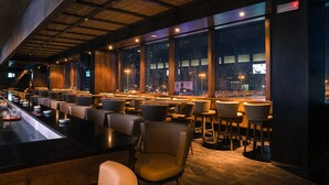 Un bar moderno con una barra larga, varias zonas de estar y grandes ventanales con vistas a la ciudad.