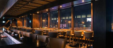 A modern bar with a long counter, multiple seating areas, and large windows offering a city view.