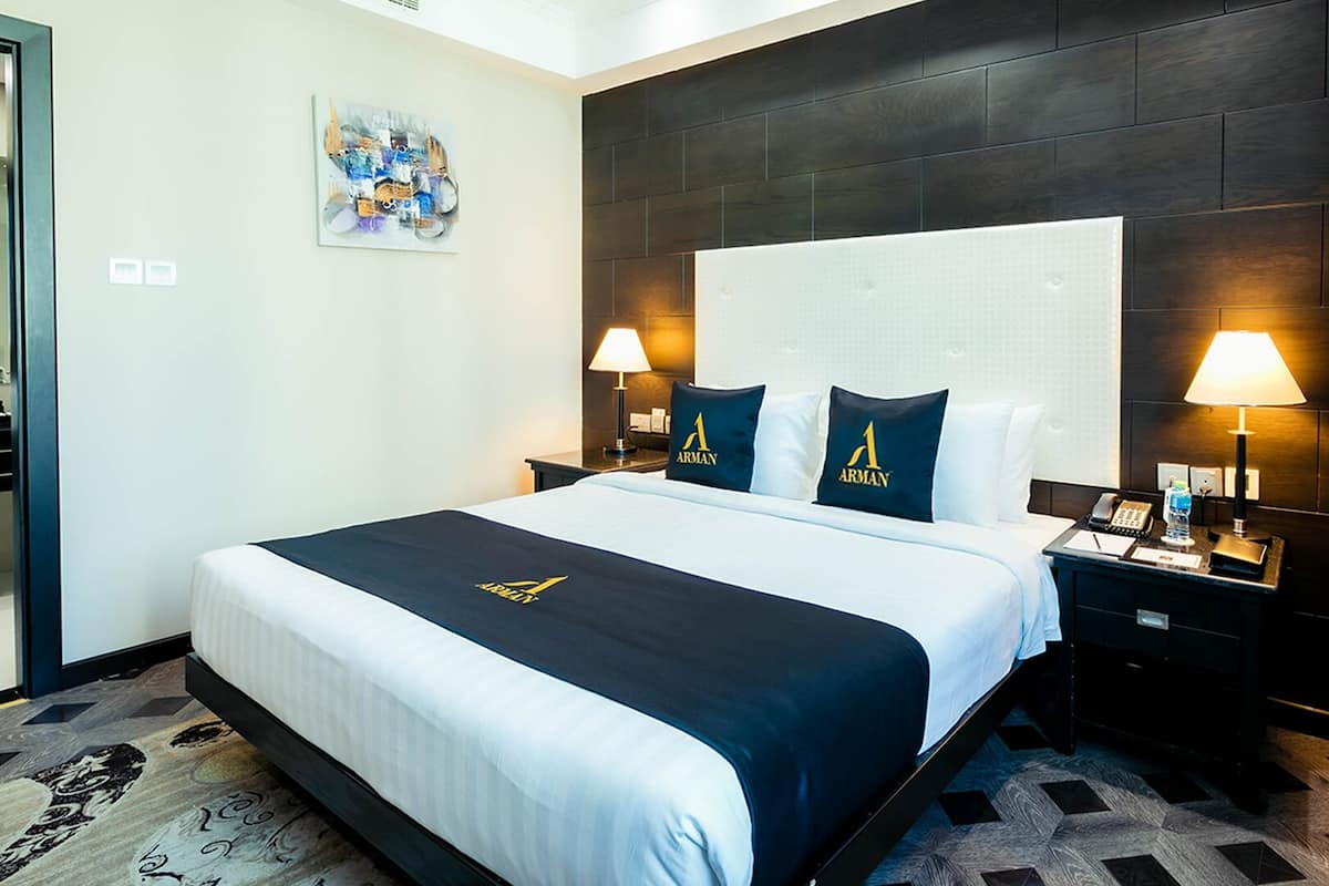 Una camera d'albergo con un letto grande, una testiera scura, due lampade ai comodini e un tavolino con un telefono e una bottiglia d'acqua.