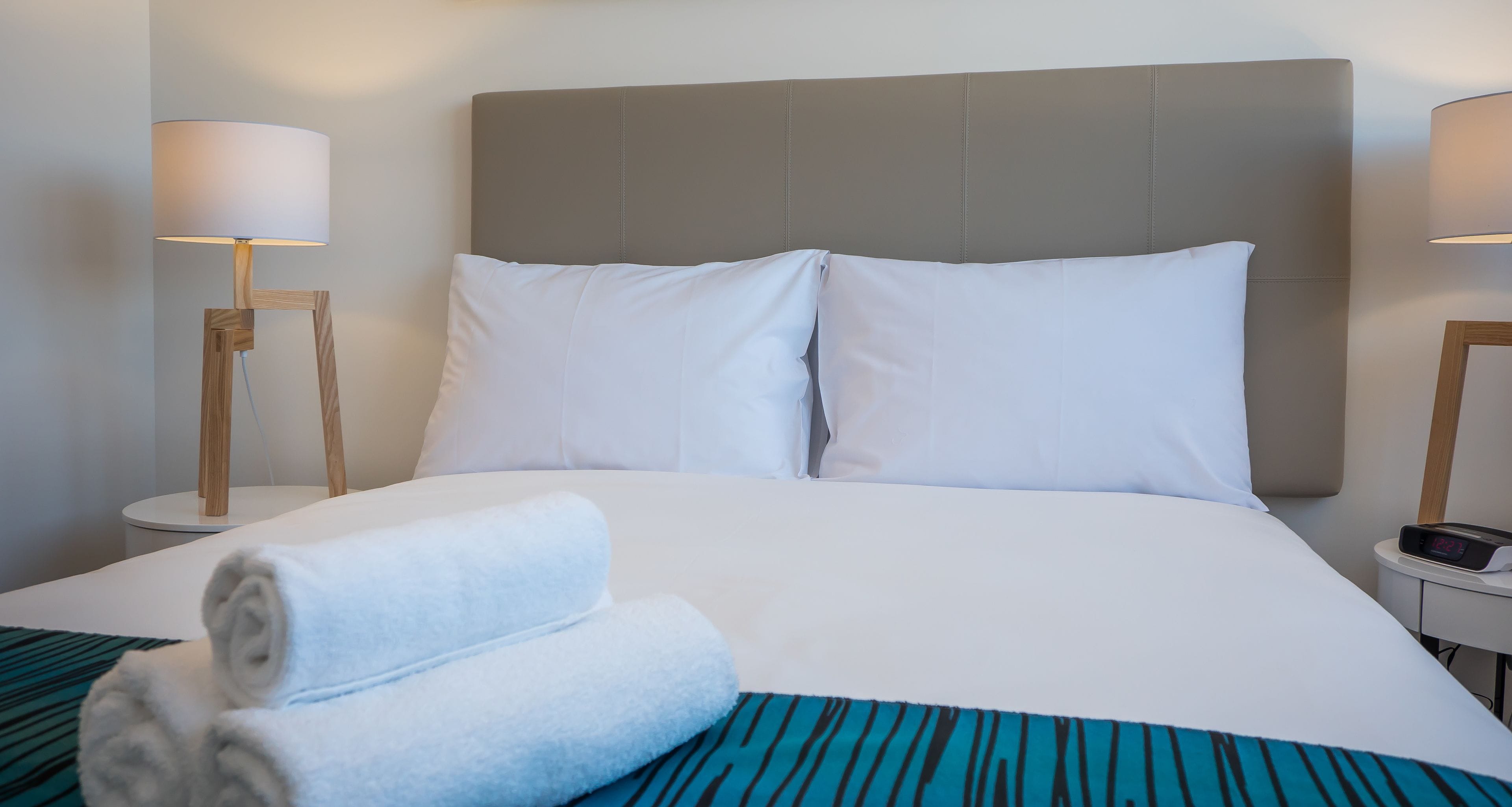 A neatly made bed with white linens, two pillows, and a folded towel. There are two bedside tables with lamps and a digital clock.