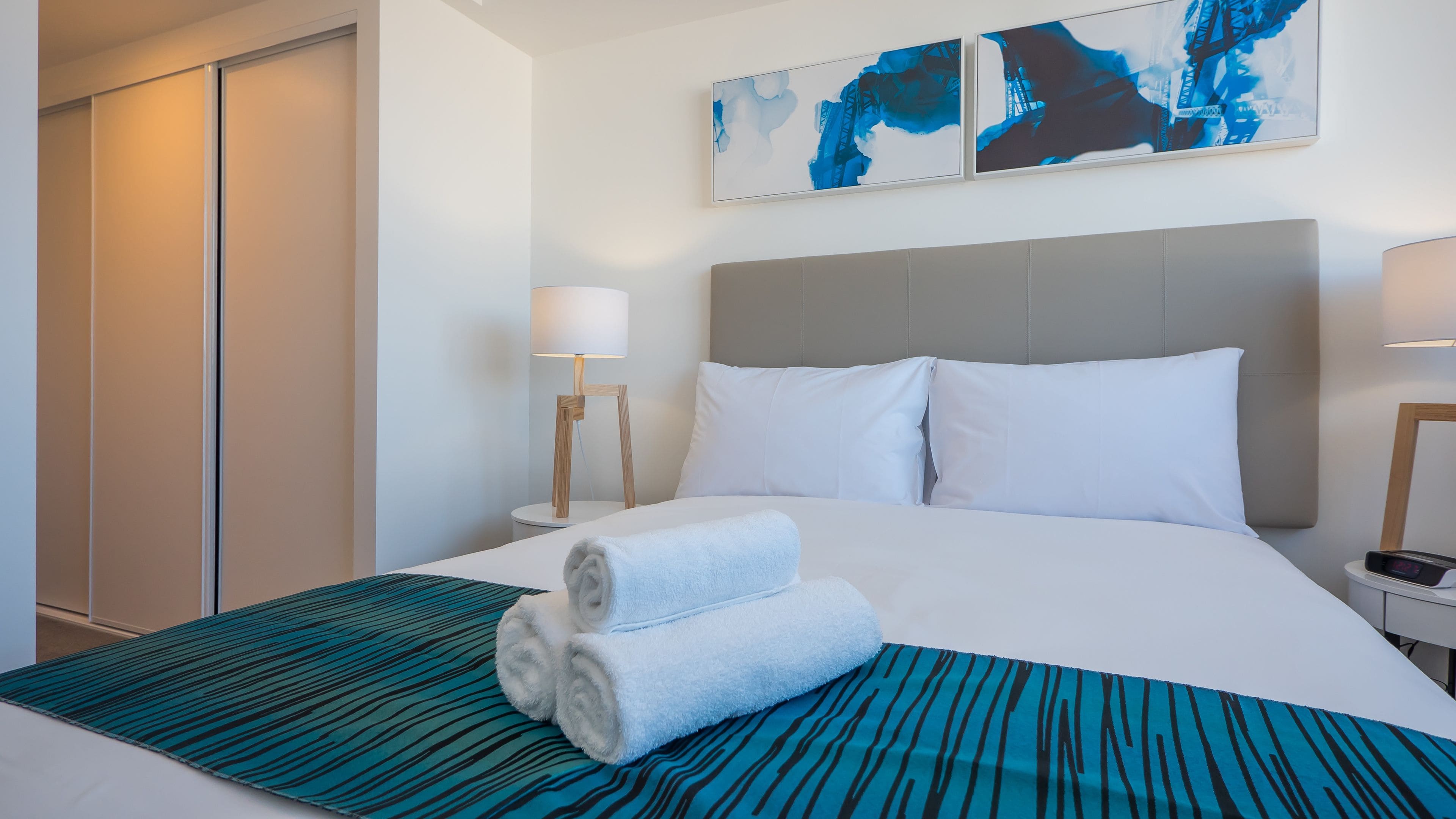 Een hotelkamer met een bed, twee kussens, een opgevouwen handdoek, een nachtkastje met een lamp en abstracte wanddecoratie.