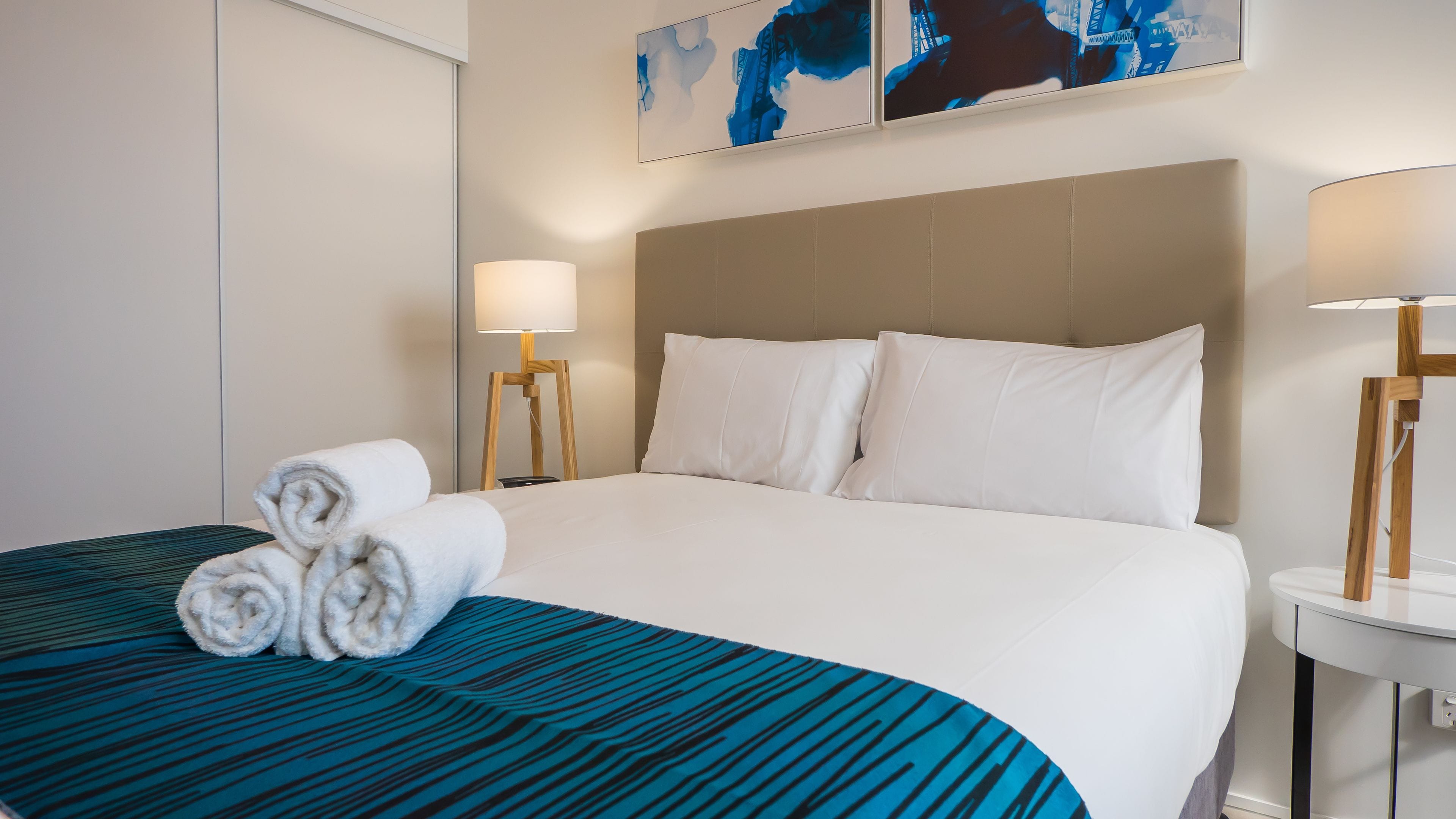 A hotel room with a neatly made bed, two bedside lamps, and a large abstract painting on the wall.