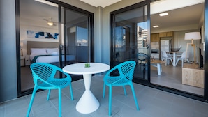 Una habitación de hotel moderna con balcón, zona de comedor y cocina visible a través de puertas de vidrio.