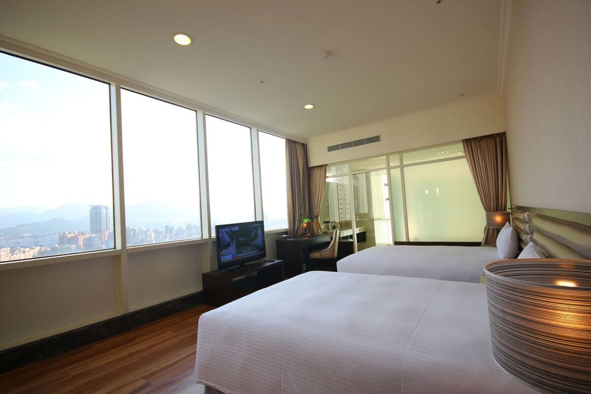 A hotel room with large windows, a flat-screen TV, a bed, a desk, and a lamp.