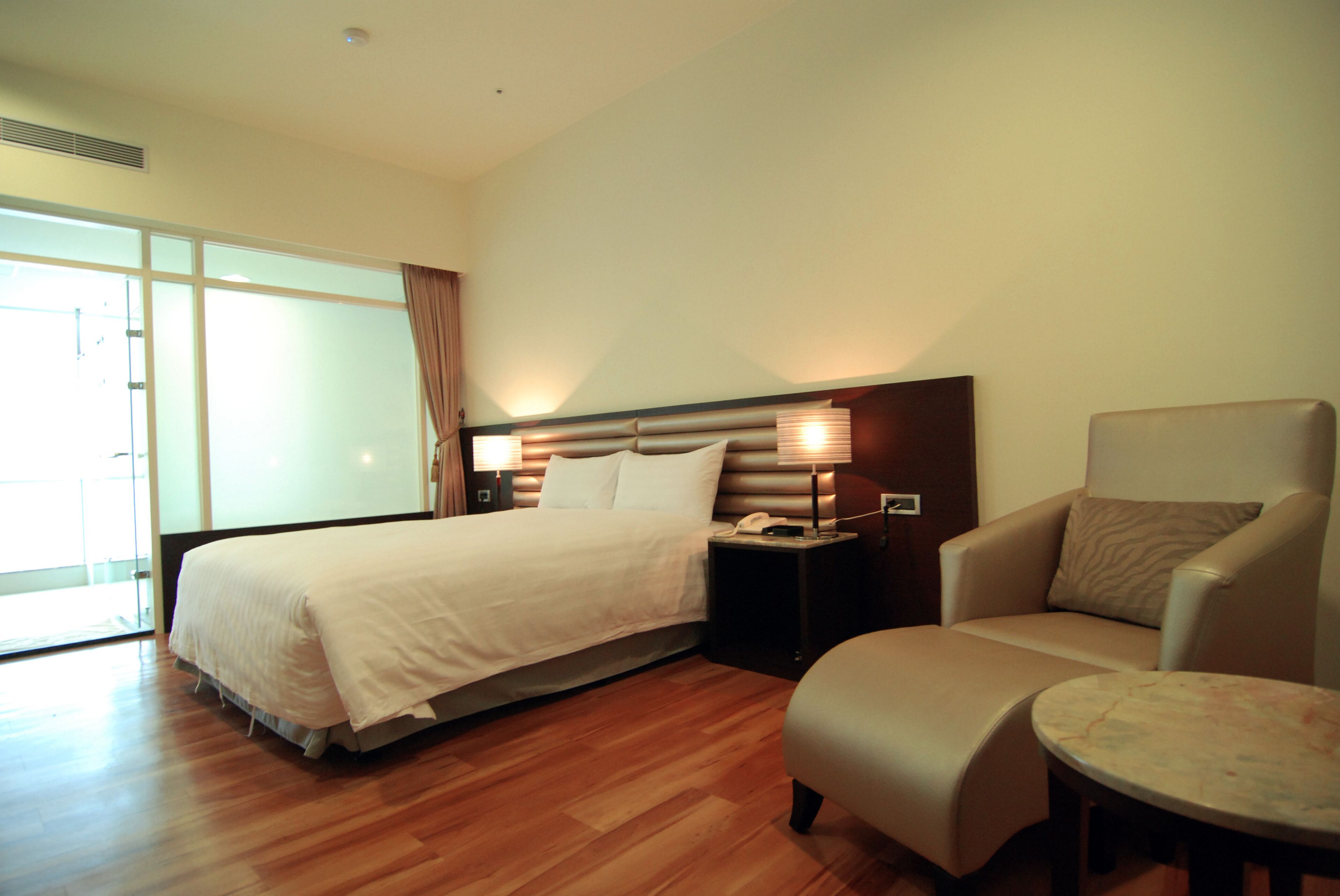 A hotel room with a large bed, a chair, a small table, and a bedside lamp.
