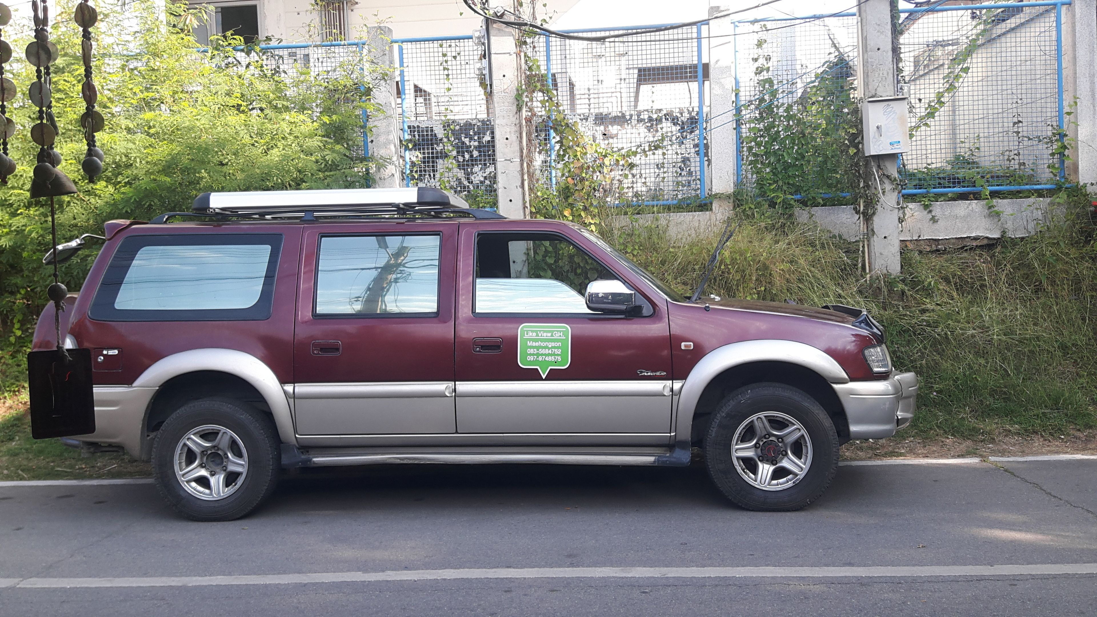 A maroon and beige SUV parked on a street with a metal fence and greenery in the background.