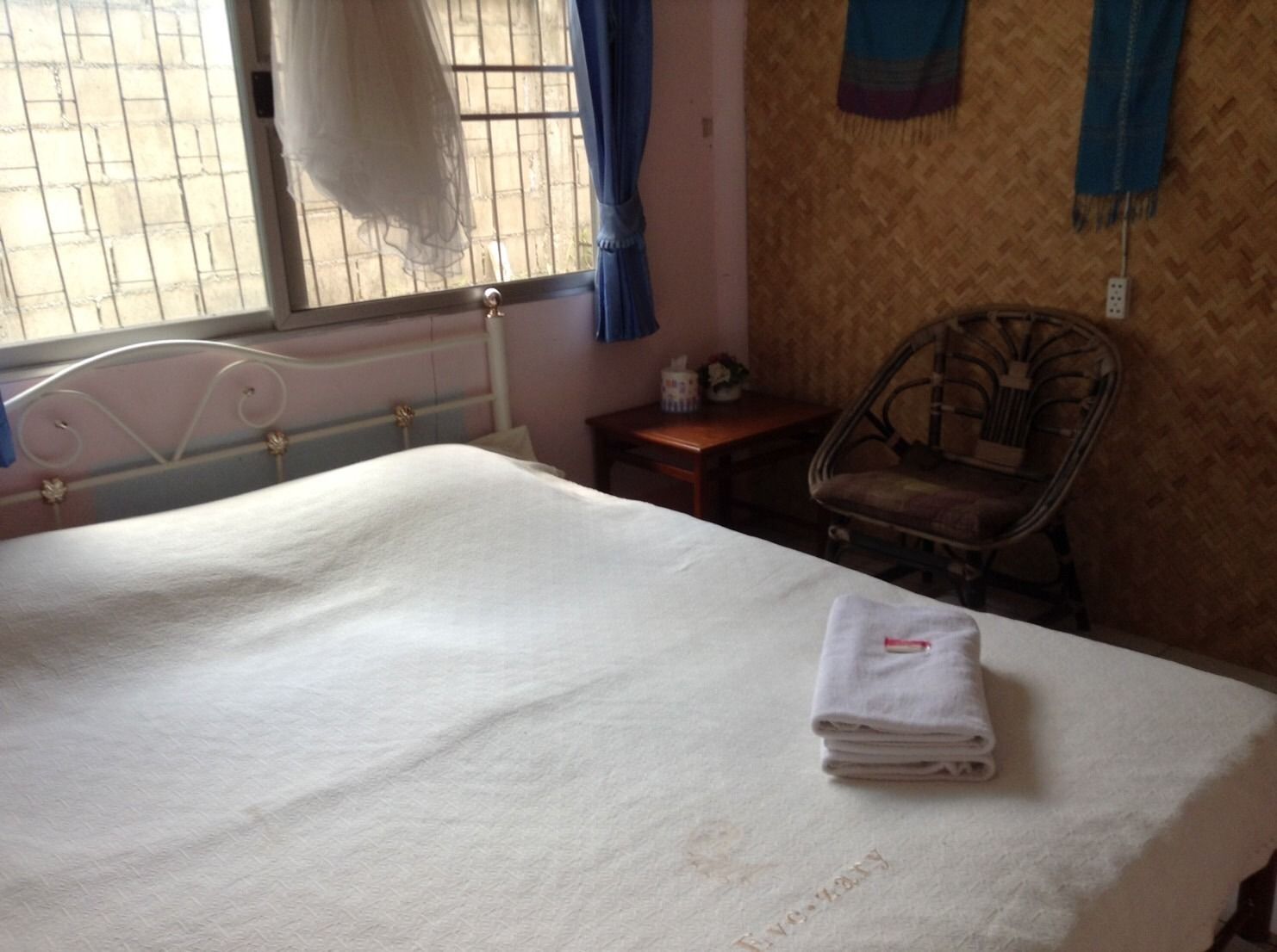 Una cama bien hecha con una toalla blanca doblada, una silla de mimbre y una mesita de noche con una lámpara.