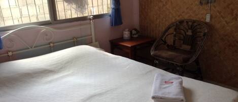 Una cama bien hecha con una toalla blanca doblada, una silla de mimbre y una mesita de noche con una lámpara.