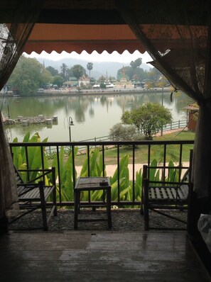 A balcony with two wooden chairs and a small table, overlooking a calm lake with buildings and trees in the background. - The Like View Guesthouse (Mae Hong Son)