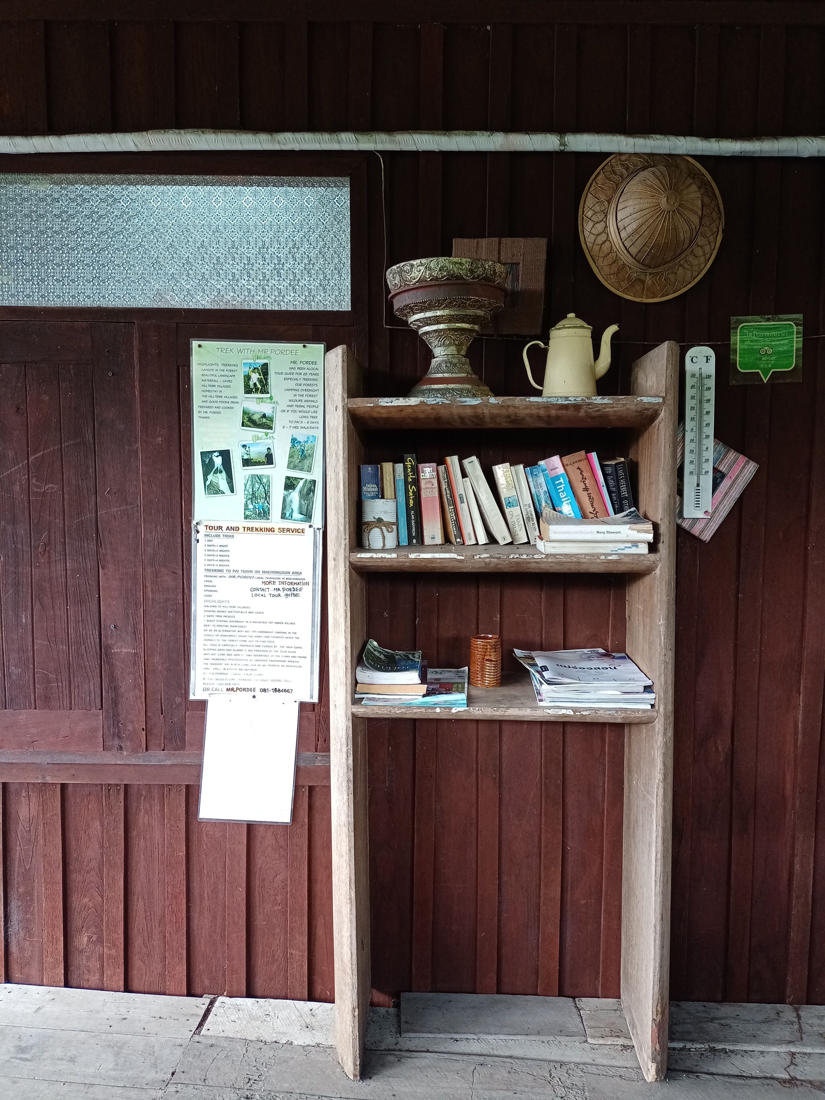 A wooden bookshelf with books and a teapot, a poster with trekking information, and a thermometer on a wall.