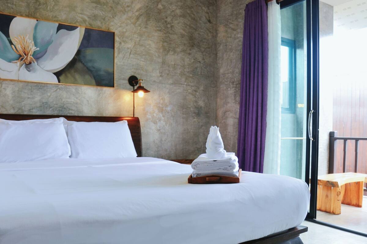 A hotel room with a large bed, a painting on the wall, a nightstand with a lamp, and a view through a glass door.