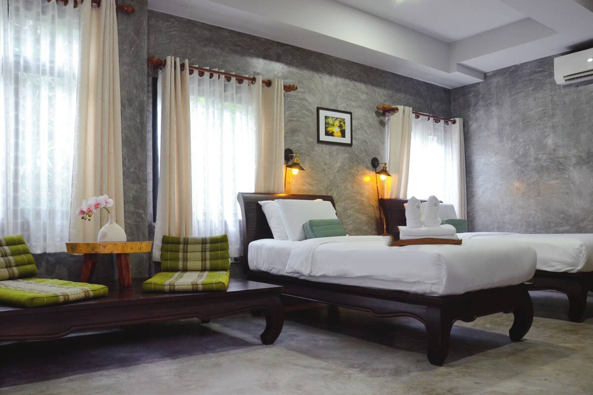A hotel room with two beds, a sofa, a small table with a vase, and two wall-mounted lamps.