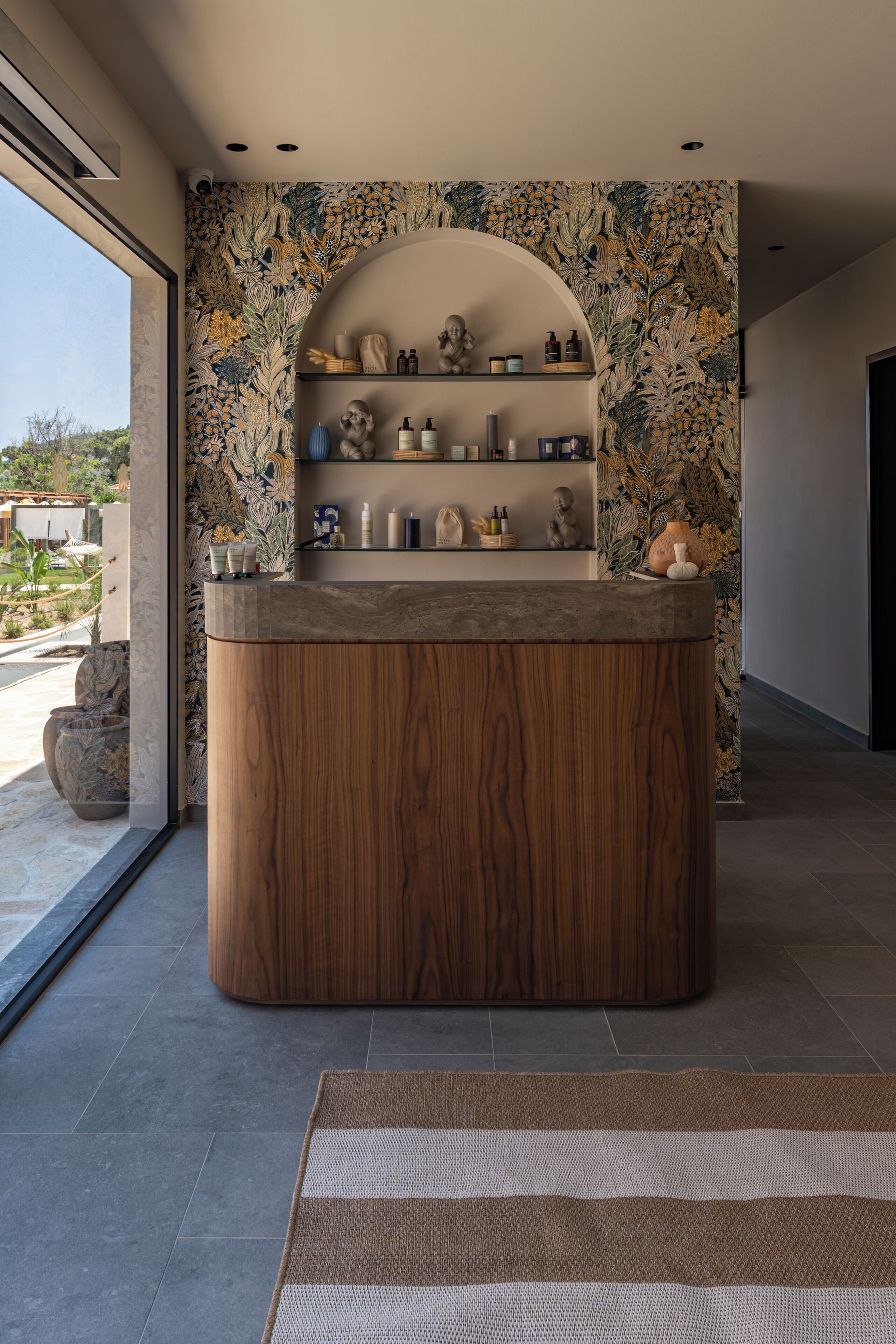 A wooden reception desk with a floral wallpaper backdrop, a large vase, and a small statue.