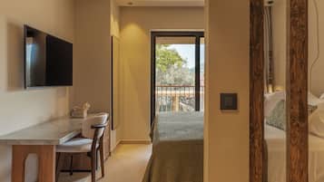 Habitación de hotel moderna con televisor de pantalla plana, zona de comedor con mesa y sillas, balcón con vistas a árboles y cama con cubrecama estampado.