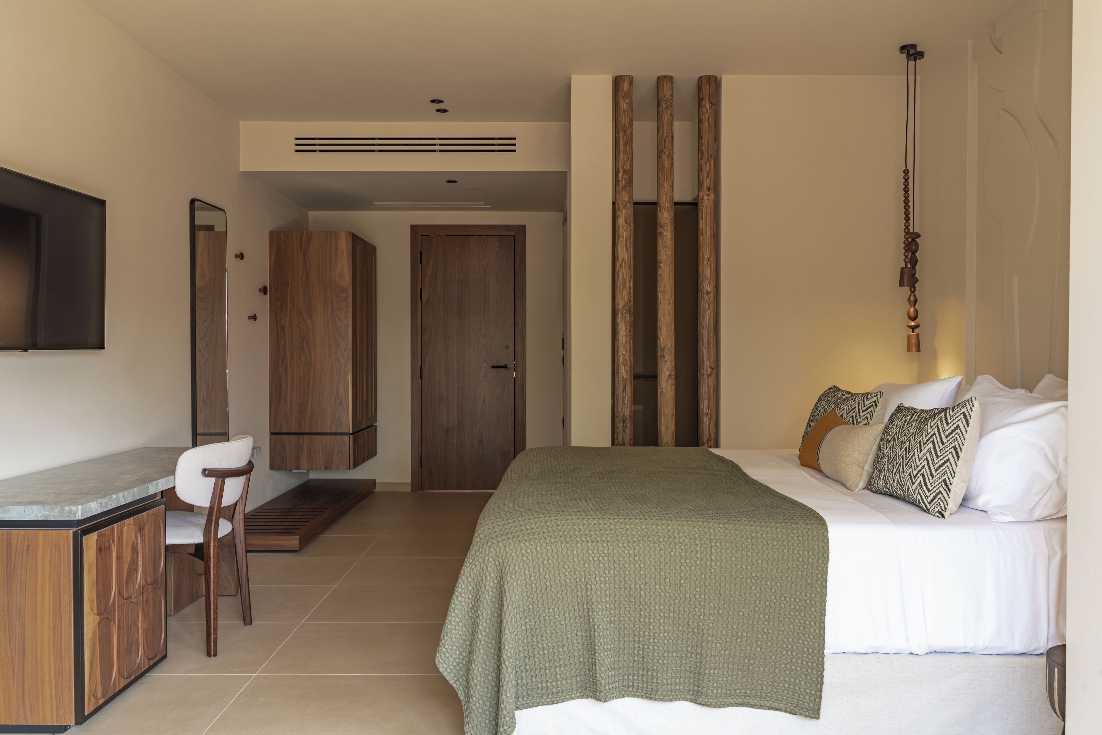A bedroom with a bed, bedside table, and a chair. There is a wooden wardrobe and a door in the background.