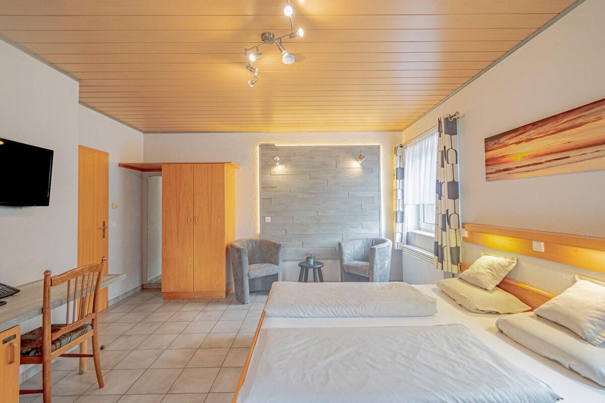 A hotel room with a bed, two chairs, a desk, a wardrobe, and a TV.