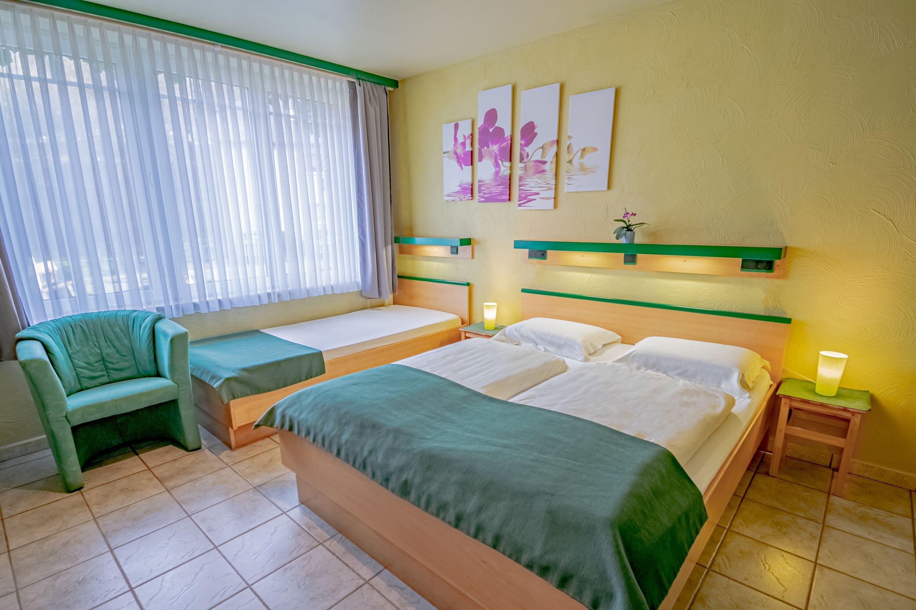 Habitación de hotel con dos camas, un sofá verde, una ventana con persianas y decoración en la pared.