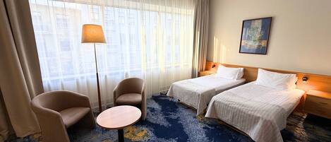 Habitación de hotel con dos camas, una mesa redonda, dos sillones, una lámpara de pie y un cuadro en la pared.