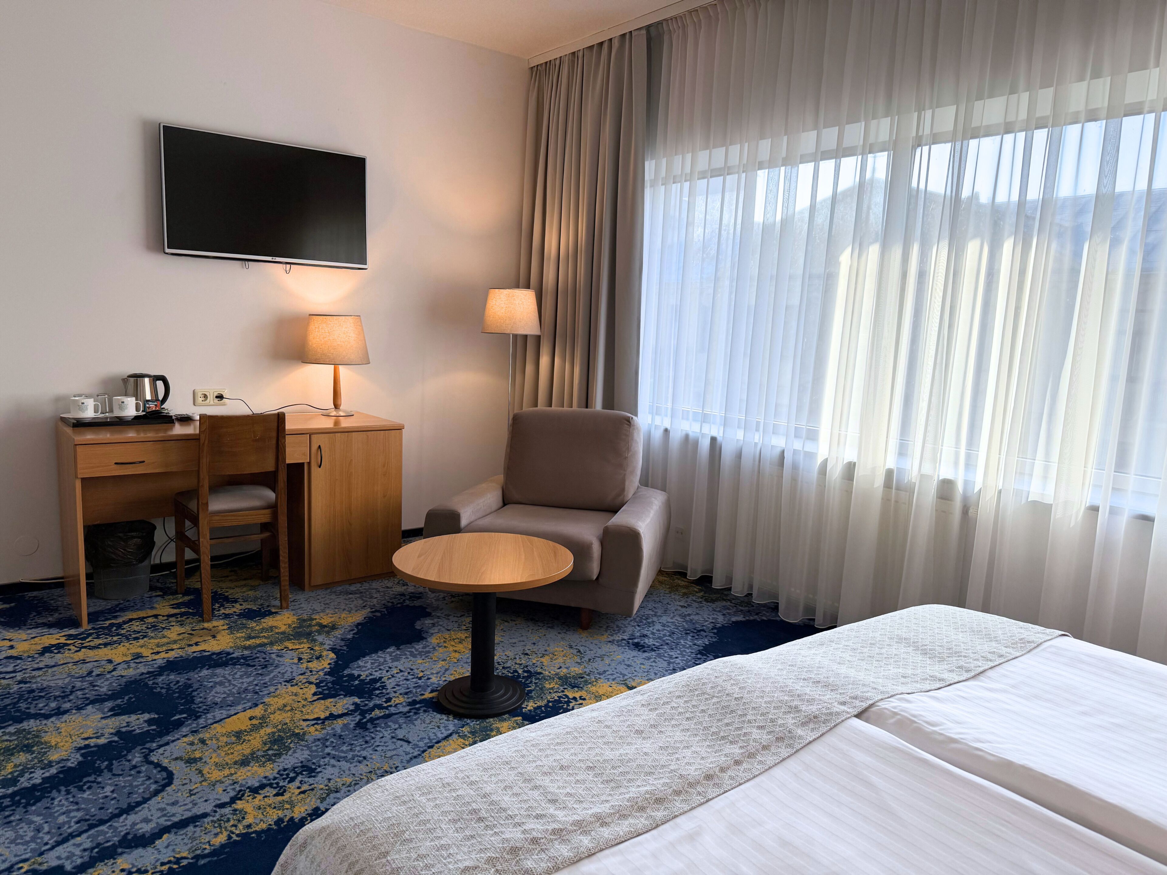 A hotel room with a bed, a desk with a chair, a small round table, a TV, and a window with sheer curtains.