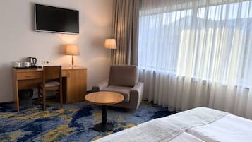 A hotel room with a bed, a desk with a chair, a small round table, a TV, and a window with sheer curtains.