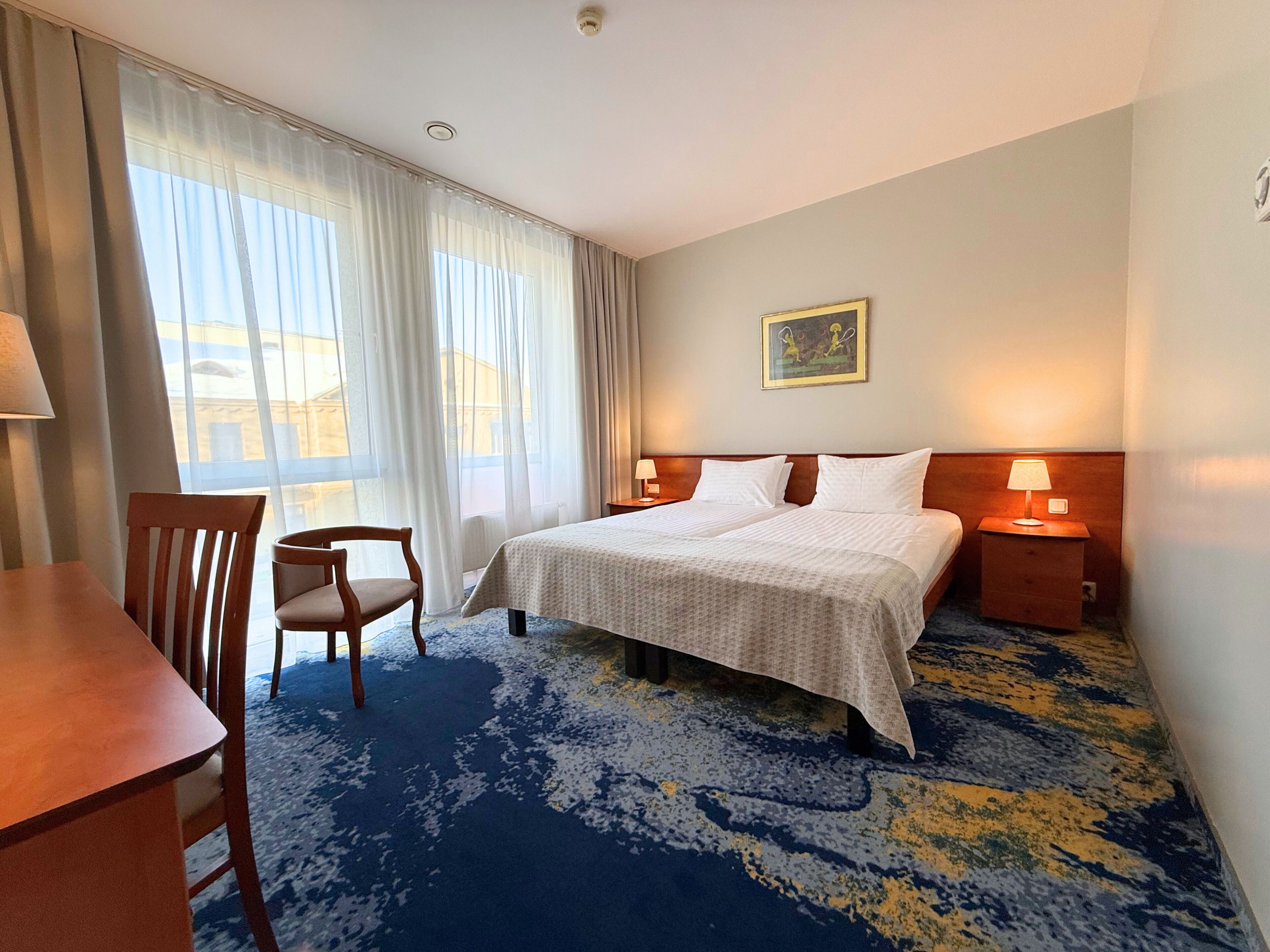 Ett hotellrum med en säng, ett skrivbord, en stol, ett nattduksbord och ett fönster med gardiner.