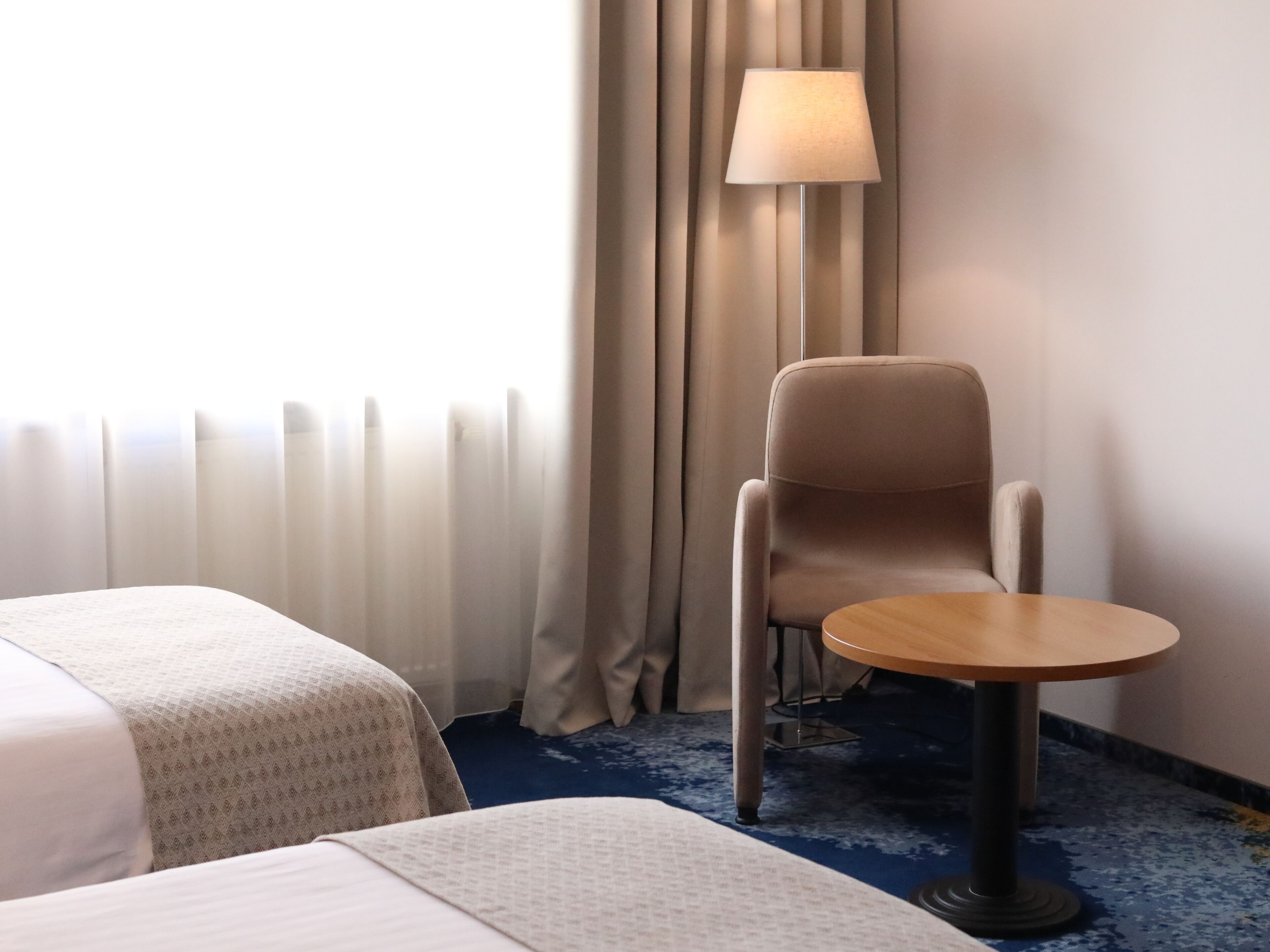 A hotel room with two beds, a chair, a small table, a lamp, and a window with curtains.