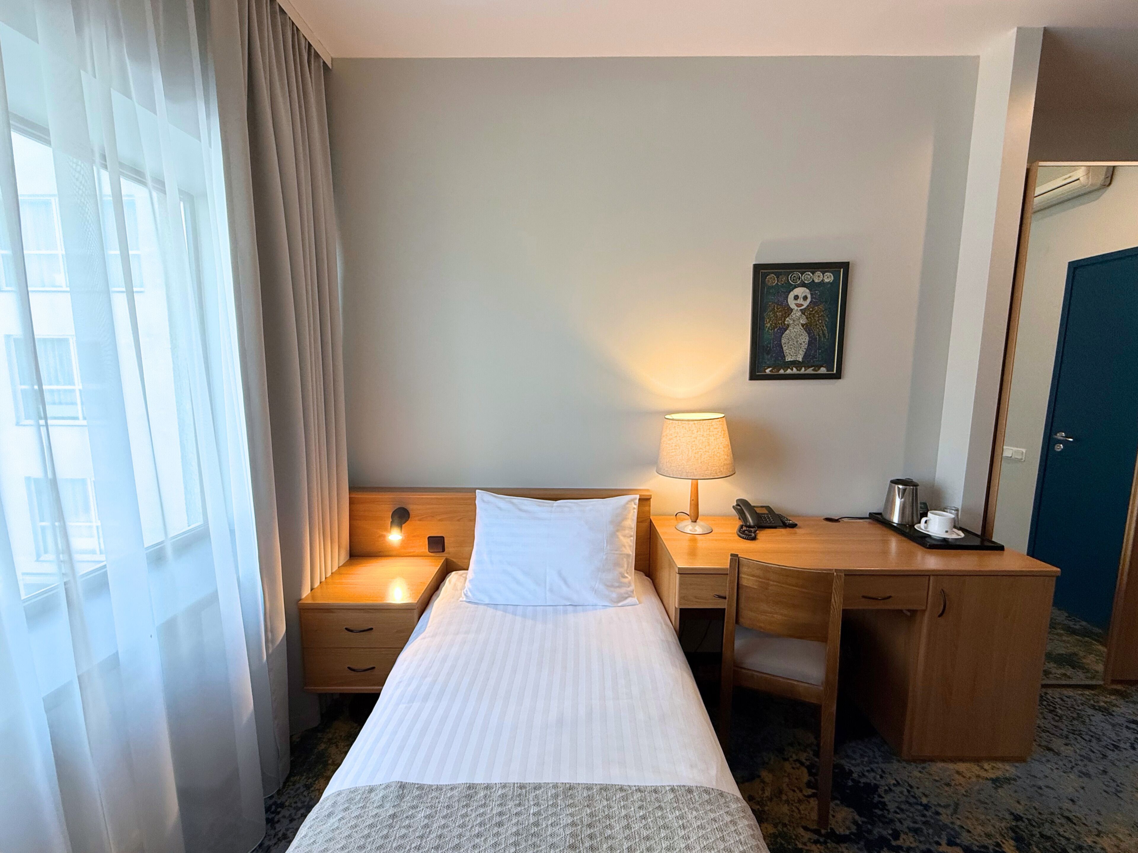 A hotel room with a bed, bedside table, lamp, desk, chair, and a framed picture on the wall.