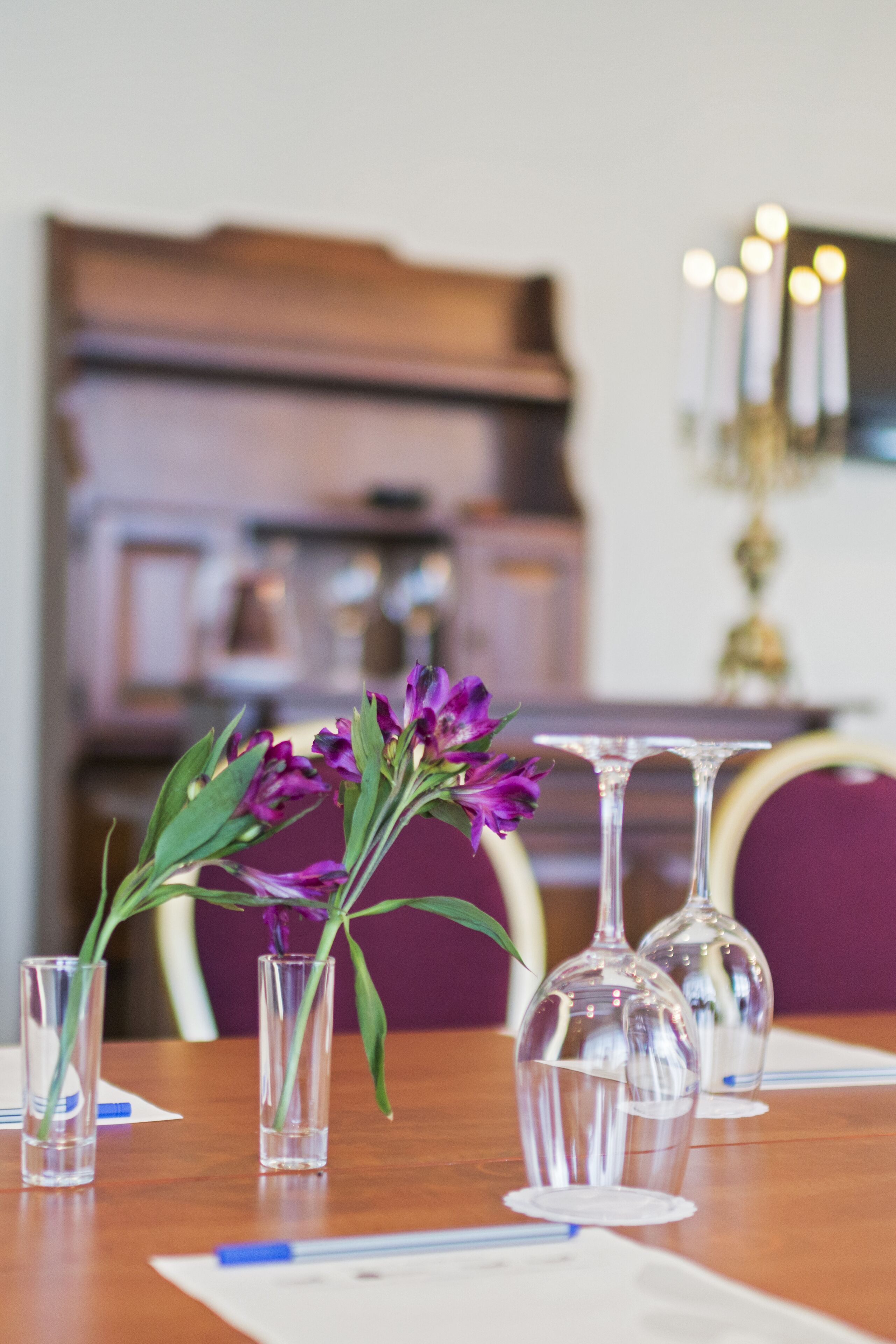 Una mesa de comedor con jarrones de vidrio transparente que contienen flores moradas, una mesa de madera y una lámpara de araña al fondo.